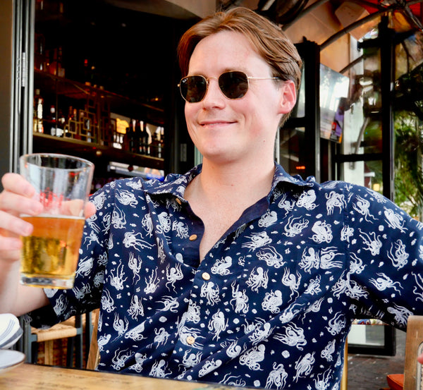 Man in a bar wearing sunglasses and a patterned shirt, holding a glass of beer.